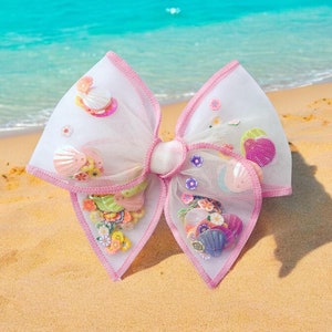 May include: A pink tulle bow with a white center and colorful embellishments. The bow is decorated with various shapes and designs, including flowers, seashells, and stars.