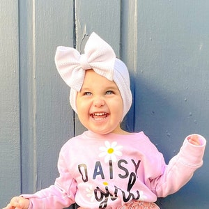 May include: A young child wearing a pink shirt with the text "Daisy girl" and a white flower graphic. The child is also wearing a white bow headband and pink floral print bloomers. The child is standing in front of a gray wall.