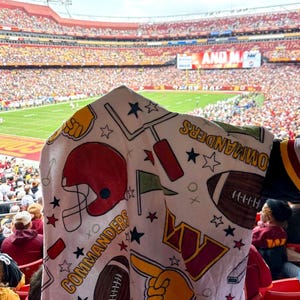 May include: A white towel with a red, yellow and black pattern featuring the Washington Commanders logo, footballs, stars and the word "Commanders".