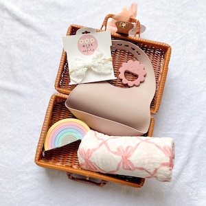 Puede incluir: Una cesta de mimbre llena de artículos para bebés, que incluyen un babero de silicona rosa con un mordedor en forma de flor, un juguete de apilar de colores arcoíris, una diadema de encaje blanco y una manta de muselina rosa y blanca. La cesta también incluye una tarjeta con el texto "WEAR wafers".