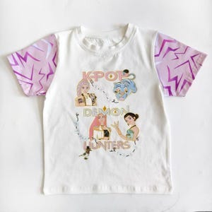 May include: White t-shirt with pink and purple lightning bolt patterned sleeves. The shirt features the text "KPOP DEMON HUNTERS" with illustrations of anime-style characters and a blue tiger.