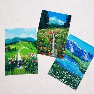 May include: Three watercolor paintings depicting different landscapes. The first painting shows a green grassy path winding through a field of wildflowers. The second painting shows a path leading through a field of wildflowers with mountains in the background. The third painting shows a mountain lake with snow-capped mountains in the background.