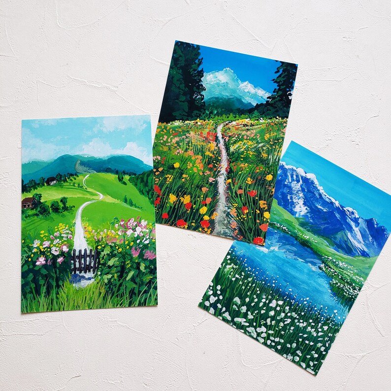 May include: Three watercolor paintings depicting different landscapes. The first painting shows a green grassy path winding through a field of wildflowers. The second painting shows a path leading through a field of wildflowers with mountains in the background. The third painting shows a mountain lake with snow-capped mountains in the background.