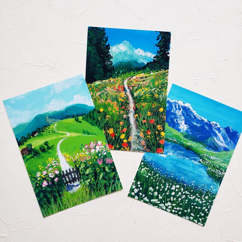 May include: Three hand-painted landscape paintings on white paper. The first painting shows a winding path through a green meadow with a small wooden fence. The second painting shows a path through a field of colorful flowers. The third painting shows a mountain range with a blue lake in the foreground.