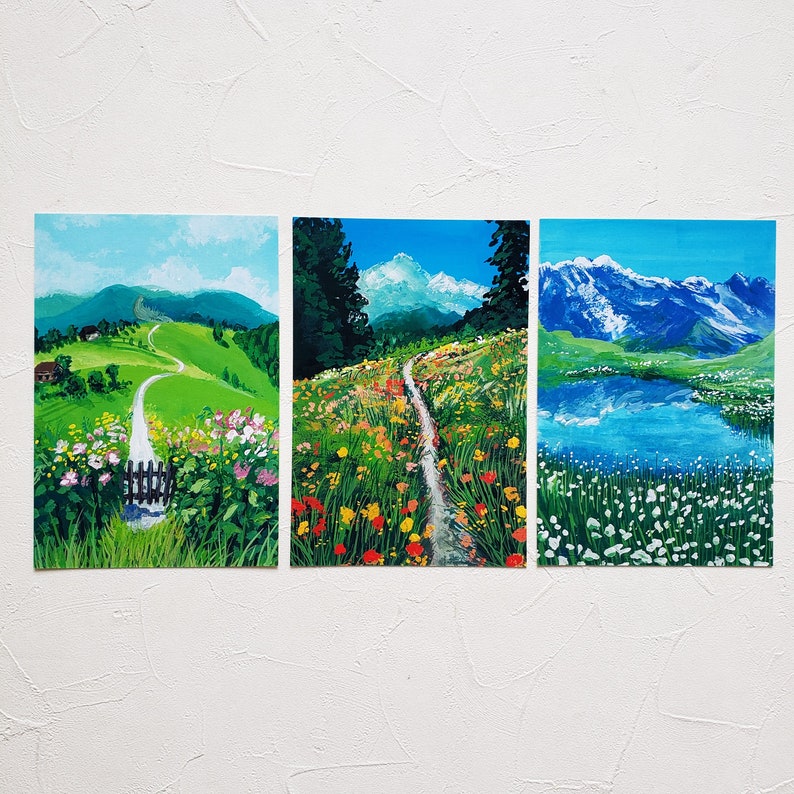 May include: Three paintings depicting different landscapes. The first painting shows a winding path leading to a small house in a green valley. The second painting shows a path through a field of wildflowers with a mountain range in the background. The third painting shows a lake with snow-capped mountains in the background.