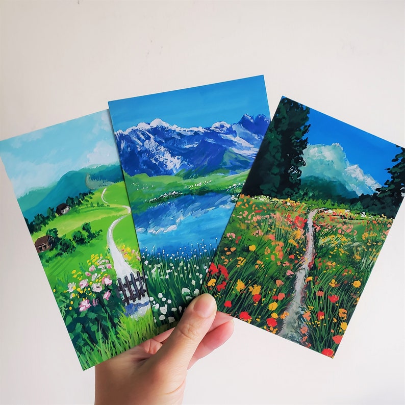 May include: Three watercolor paintings of landscapes. The first painting shows a green field with a winding path leading to a small house. The second painting shows a mountain range with a lake in the foreground. The third painting shows a field of wildflowers with a path leading through the center.