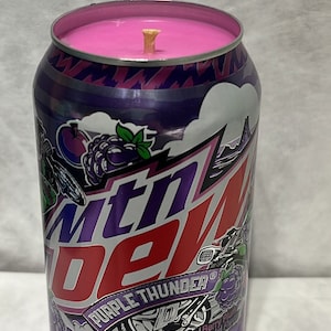 May include: A purple and silver can candle with a pink wax top and a wooden wick. The can has the Mountain Dew logo and the text "Purple Thunder" with a blast of berry plum, natural and artificial flavors.
