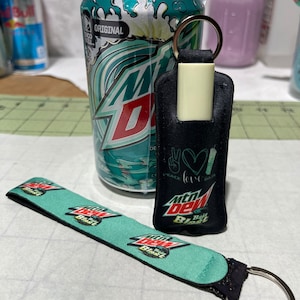 May include: A can of Mountain Dew Baja Blast soda, a black keychain with the Mountain Dew logo, and a matching teal wrist strap. The can is wrapped in plastic. The keychain has a silver ring.