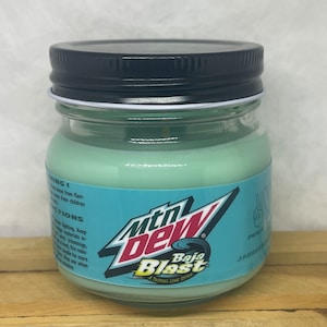 May include: A green glass jar candle with a black lid. The jar has a label with the Mountain Dew Baja Blast logo and the text "A Tropical Lime Storm".
