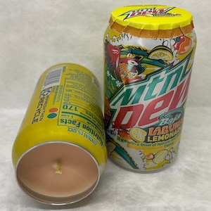 May include: Two yellow and green Mountain Dew Baja Laguna Lemonade cans. One can is a candle with a peach-colored wax. The can has the text "REFRESH THEN RECYCLE" on the side. The other can has the text "DEW with a Blast of Natural Mango Lemonade Flavor" on the side.