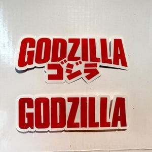May include: Two red and white stickers with the word "GODZILLA" in bold, capitalized letters. One sticker includes Japanese characters. The stickers are rectangular with rounded corners.