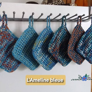 Pode incluir: Sete pegadores de panela de crochê em tons de azul, marrom e branco pendurados em um rack de metal. Os pegadores de panela são quadrados e têm um design texturizado de crochê. O texto "L'Amelina bleue" é visível na parte inferior da imagem.