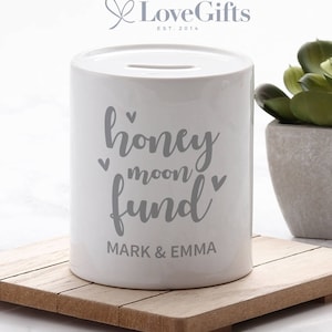 May include: White ceramic money box with a slot on the top. It has the text "honeymoon fund" with hearts and the names "Mark & Emma" printed on the front.