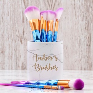 May include: A white marble makeup brush holder with the text "Amber's Brushes" in gold lettering. The holder is filled with a set of makeup brushes with blue and pink handles.