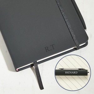 May include: A dark grey notebook with the initials "R.T" embossed on the cover. A black pen is attached to the notebook. The pen has the name "RICHARD" printed on it. A black elastic band and a black ribbon bookmark are also visible.