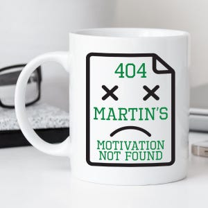 May include: A white ceramic mug featuring a black outlined document icon with green text that reads 404, MARTIN'S, and MOTIVATION NOT FOUND. The design includes two black X symbols for eyes and a curved black line representing a sad mouth.