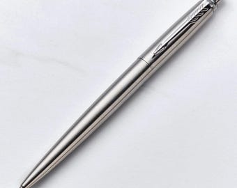 May include: A silver-colored ballpoint pen with a sleek, metallic finish. The pen features a pointed tip and a clip with an arrow design. The pen is displayed on a white background.