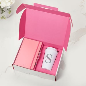 May include: A pink gift box containing a coral notebook with the initials N.T. embossed on the cover, a matching pink pen, and a white tumbler featuring a large grey letter S and the name Sophie in pink script lettering.