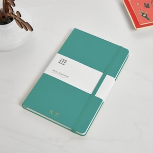 May include: Teal Moleskine Classic notebook with a white label that says "Moleskine Classic Notebook" and a gold elastic closure. The notebook has the initials "K.E." embossed in gold on the front cover.