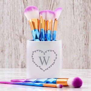 May include: White ceramic makeup brush holder with a heart design and the letter "W" in the centre. The holder is filled with pink and blue makeup brushes.
