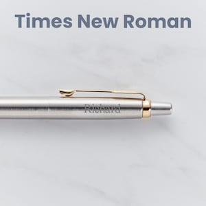 May include: A silver pen with a gold clip and a gold band at the end. The pen is engraved with the name "Richard" in a Times New Roman font.