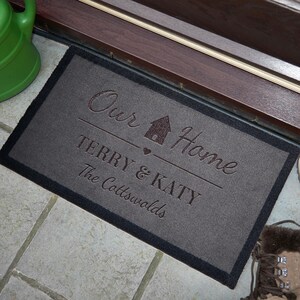May include: A grey doormat with black edging features the text "Our Home" with a house icon and heart. Below the text is "Terry & Katy" and "The Cotswolds".