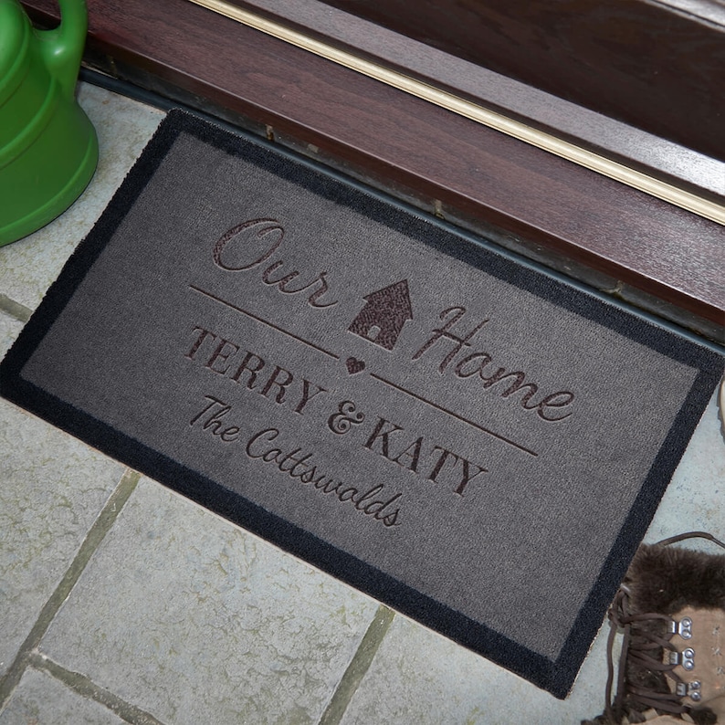 May include: A grey doormat with black edging features the text "Our Home" with a house icon and heart. Below the text is "Terry & Katy" and "The Cotswolds".