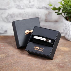 May include: A silver magnetic bracelet with the initials "D.M." engraved on it. The bracelet is in a black box with the brand name "magrelief MAGNETIC PRODUCTS" printed on it.
