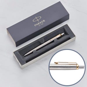 May include: A silver Parker pen with a gold trim and the inscription "Richard" on the side. The pen is in a white box with a black lid that has the Parker logo and the text "Est. 1888".