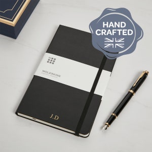 May include: A black Moleskine notebook with a white band and gold initials "J.D". A black and gold pen is next to the notebook. The image includes a "Hand Crafted" seal with a Union Jack.