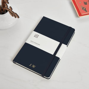 May include: A dark blue Moleskine notebook with a white elastic band and a white label that says "Moleskine Legendary Notebooks". The notebook has a gold embossed "J.W" on the front cover.