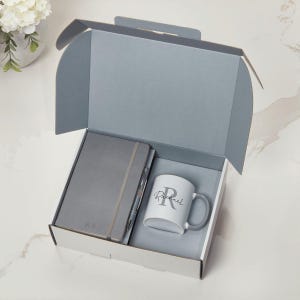 May include: A grey gift box containing a grey notebook, pen, and a white mug with a grey handle. The notebook has the initials "R.S." and the mug has the name "Rachael" printed on it. The box is open, revealing the contents.