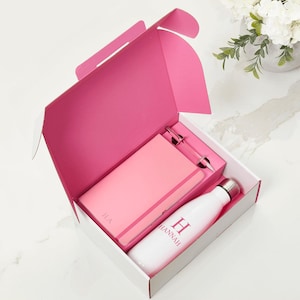 May include: A gift set in a pink and white box containing a pink notebook with the initials H.A., a pink pen, and a white water bottle with a silver cap featuring the name HANNAH and a large letter H printed in pink on the front of the bottle.
