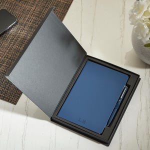 May include: A dark blue notebook with the initials L.H. embossed on the cover, paired with a matching blue pen, both presented inside a black rectangular gift box with a hinged lid. The set is arranged neatly within a custom-fitted foam insert.