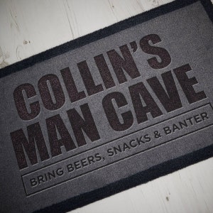May include: A gray doormat with black text that reads "COLLIN'S MAN CAVE BRING BEERS, SNACKS & BANTER".