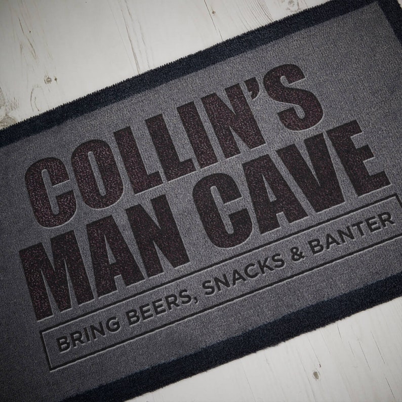 May include: A gray doormat with black text that reads "COLLIN'S MAN CAVE BRING BEERS, SNACKS & BANTER".