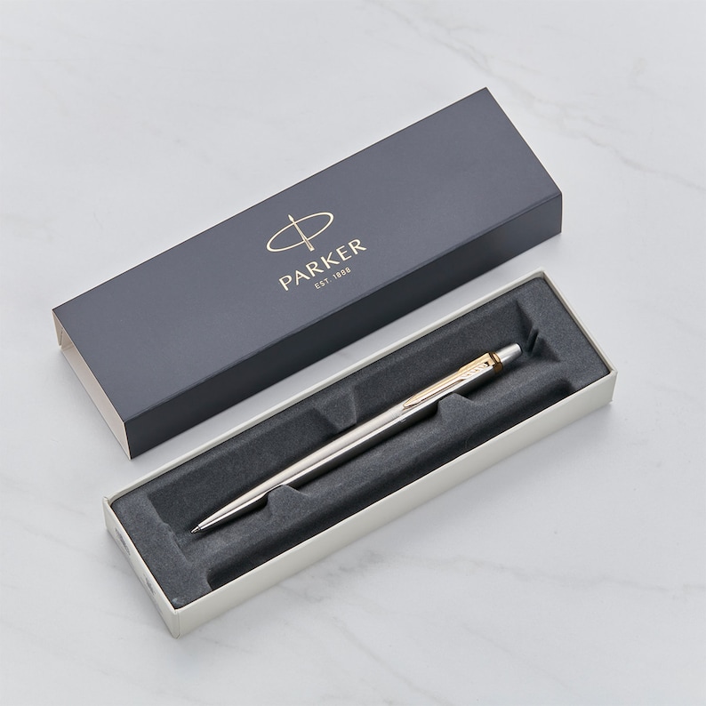 May include: A silver Parker pen with a gold trim in a white box with a black interior. The box has a black lid with the Parker logo and the text "EST. 1888".