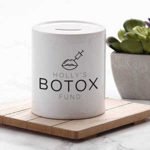 May include: White ceramic money box with a black line drawing of lips with a needle injecting them. The text "HOLLY'S BOTOX FUND" is printed below the image.