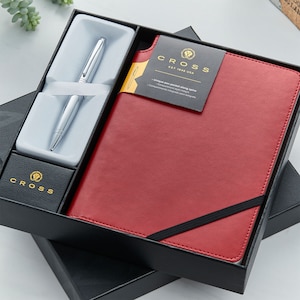 May include: A black gift box containing a red leather journal and a silver pen. The journal has a black elastic band and the text "CROSS EST. 1846 USA" on the front. The pen is in a white box with the CROSS logo.