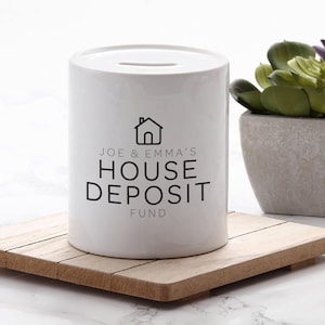 May include: A white ceramic money bank with a slot on top. The bank has black text that reads "JOE & EMMA'S HOUSE DEPOSIT FUND" with a house icon. The bank sits on a wooden coaster.