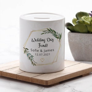 May include: White ceramic money box with a gold geometric frame and green leafy branches. The text reads "Wedding Day Fund! Sofie & James 12.07.2021" with a small gold heart.