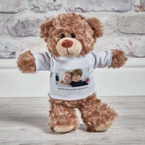 May include: A brown plush teddy bear wearing a white t-shirt. The shirt features a printed photograph of two children and the text "HAPPY BIRTHDAY CHARLIE X" printed in black ink below the image. The bear has dark eyes and a stitched nose.
