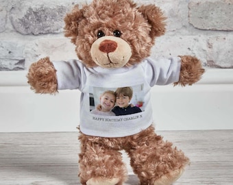 May include: A brown plush teddy bear wearing a white t-shirt. The shirt features a printed photograph of two children and the text "HAPPY BIRTHDAY CHARLIE X" printed in black ink below the image. The bear has dark eyes and a stitched nose.