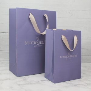 May include: Two purple gift bags with white satin handles. The larger bag has the text "Boutique Gifts Est. 2014" printed on the front. The smaller bag has the same text but with a slightly different font.