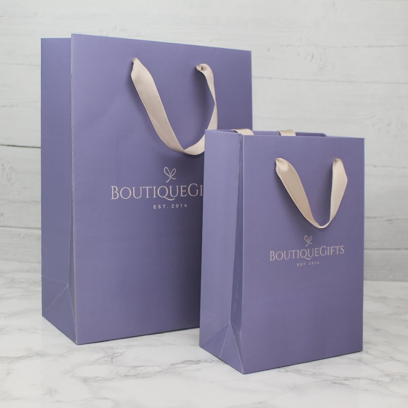May include: Two purple gift bags with white satin handles. The larger bag has the text "Boutique Gifts Est. 2014" printed on the front. The smaller bag has the same text but with a slightly different font.