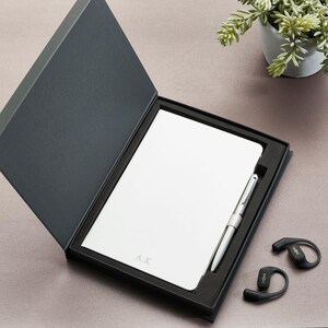May include: A white notebook with the initials A.K. embossed on the cover, presented in a black hinged gift box alongside a silver pen. Two black wireless ear hooks are positioned outside the box, featuring the brand name Shokz on the side.