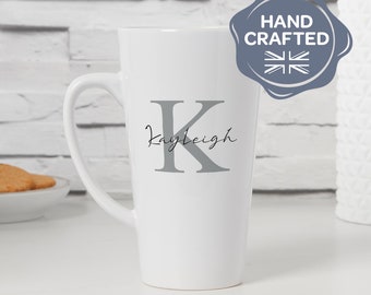 May include: White ceramic mug with a tall, slender design. The mug has a gray letter "K" and the name "Kayleigh" written in cursive script. The mug is hand-crafted and has a seal that says "Hand Crafted".