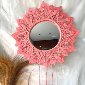 May include: A round, pink macrame mirror with a sunburst design. The mirror has a circular glass centre, surrounded by intricate knotted patterns and small wooden beads. Three pink tassels hang from the bottom.