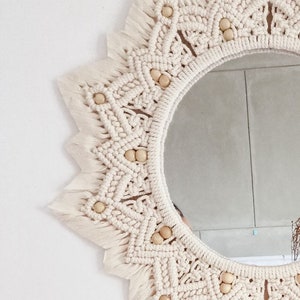 May include: A white macrame mirror with a circular shape and wooden beads around the edge. The mirror has a fringe border.