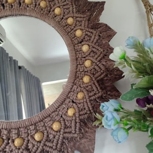 Op de afbeelding: Een bruine macramé spiegel met een ronde lijst en houten kralen. De spiegel heeft een franje rand en hangt aan een witte muur.